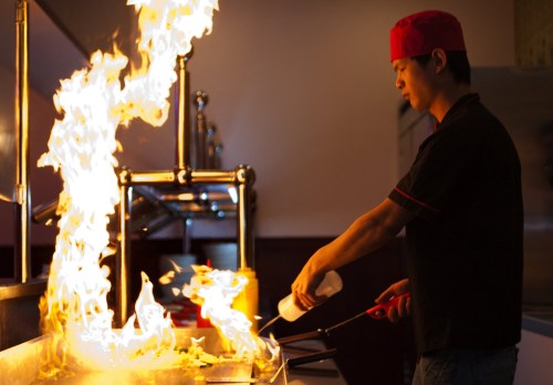 A chef in a red hat ignites flames while cooking in a restaurant kitchen, showcasing a dramatic cooking technique.