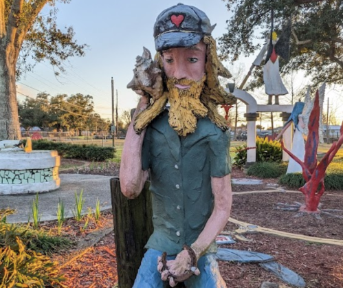 A colorful wooden sculpture of a man with a beard, wearing a cap, sitting with a shell on his shoulder in a park setting.