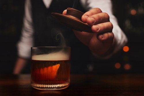 A bartender places a wooden lid over a glass of cocktail, with smoke and an orange twist visible.