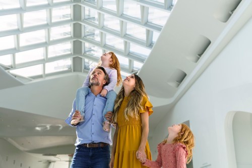 A family of four looks up at a skylight, with a father holding a child on his shoulders and two girls beside him.