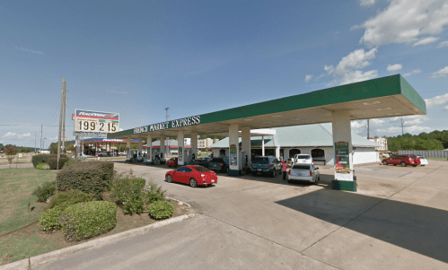 A gas station with multiple pumps, cars parked, and a sign displaying fuel prices under a clear blue sky.