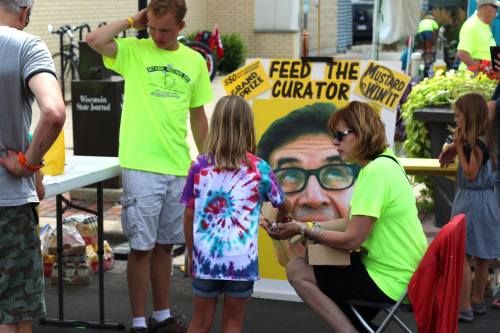 A child in a tie-dye shirt interacts with adults in neon shirts at a fair, with a humorous sign in the background.