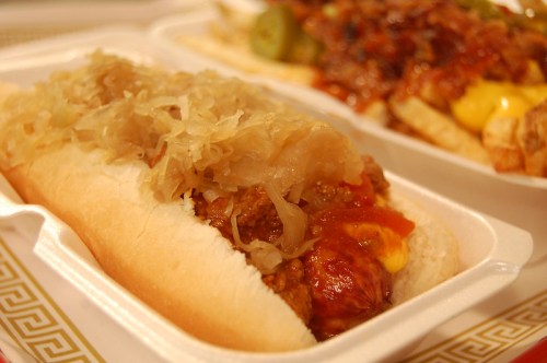 A close-up of a hot dog topped with sauerkraut and chili, served in a white takeout container.