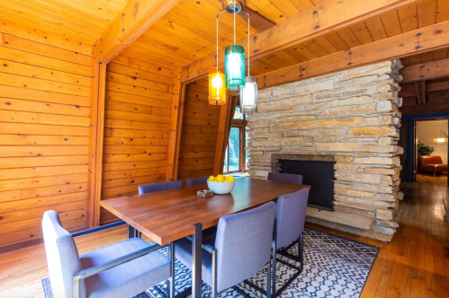 Cozy dining area with a wooden ceiling, stone fireplace, and a table set for six, illuminated by colorful pendant lights.