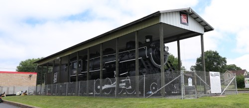 A vintage steam locomotive displayed under a shelter, surrounded by a fence, with a cloudy sky in the background.