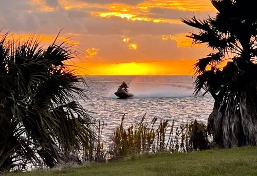 A person rides a jet ski on water at sunset, framed by palm trees and vibrant orange and yellow skies.