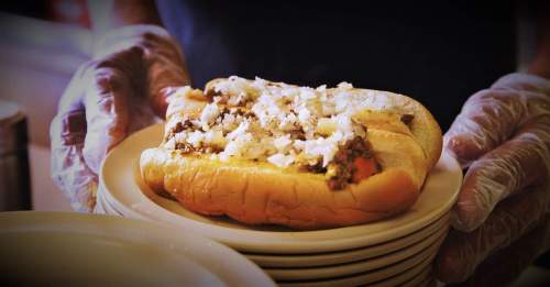 A person holding a hot dog topped with chopped onions, served on a plate, with additional plates stacked in the background.