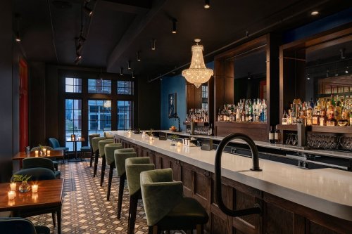 A stylish bar with a long white countertop, green chairs, and a chandelier, featuring shelves of liquor in a dimly lit setting.