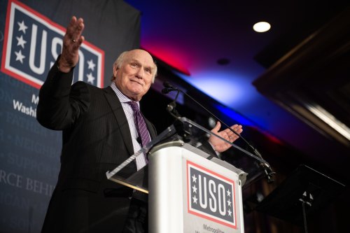 A speaker gestures while addressing an audience at a USO event, with a backdrop featuring the USO logo.