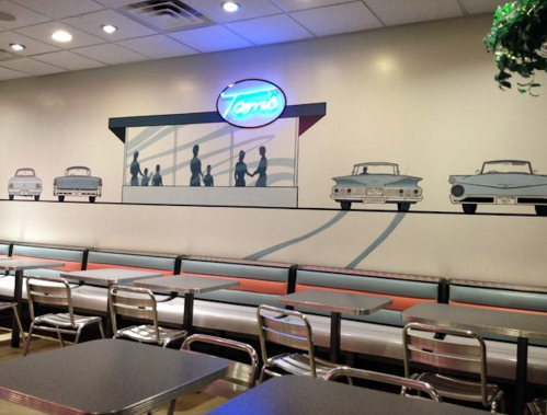 A retro diner interior with a mural of classic cars and people, featuring a neon sign that reads "Tom's."