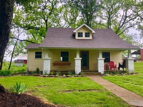 A charming yellow cottage with a front porch, surrounded by greenery and trees, on a sunny day.