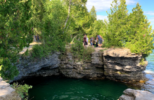 A rocky cliff surrounded by greenery, with people gathered near a deep, clear green pool of water below.