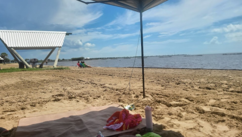 A sandy beach scene with a view of the water, a shaded area, and distant figures enjoying the outdoors.