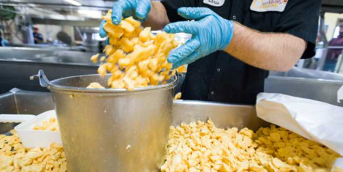 A worker in gloves pours orange snack pieces from a container into a bucket in a food processing facility.