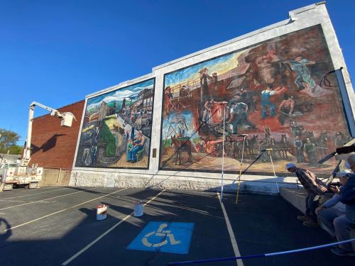 Two large murals on a building wall, depicting various scenes of labor and community life, with a blue sky above.