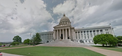 A large stone building with a dome, steps leading up, and green lawns under a cloudy sky.