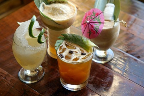 A variety of colorful cocktails with garnishes, including umbrellas and fruit, on a wooden table.