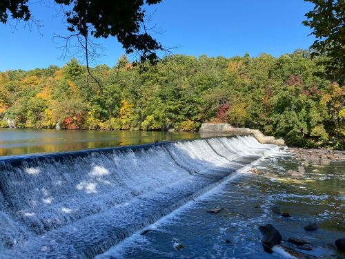A serene waterfall cascades over a dam, surrounded by vibrant autumn foliage and a clear blue sky.