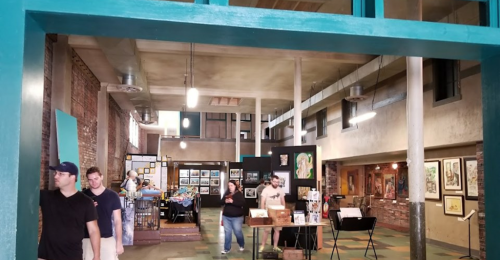 Interior of an art gallery with people browsing artwork, featuring brick walls and colorful displays.