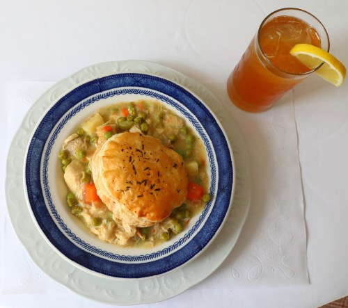 A plate of chicken pot pie with vegetables and a flaky biscuit, accompanied by a glass of iced tea with a lemon slice.
