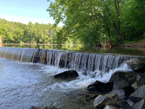 A serene waterfall cascades over rocks into a calm river, surrounded by lush green trees and a clear blue sky.