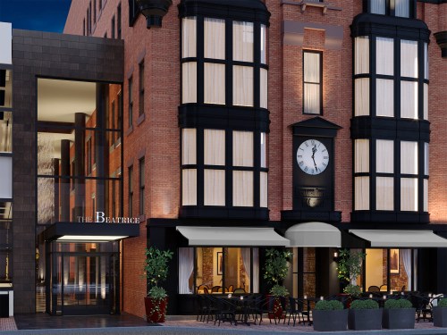 Exterior view of a brick hotel with large windows, a clock, and outdoor seating under awnings. Evening setting.
