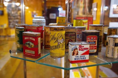 A display of vintage mustard cans in various designs and colors, arranged on a glass shelf.