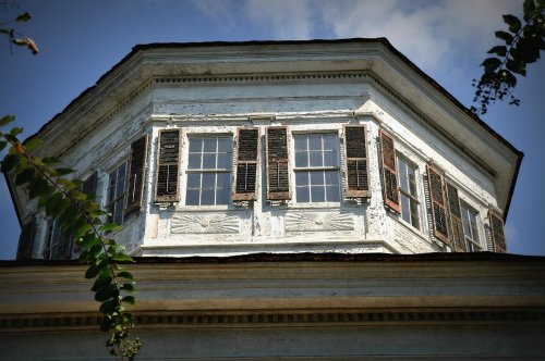 Close-up of an octagonal building with weathered white paint and multiple windows, framed by green foliage.