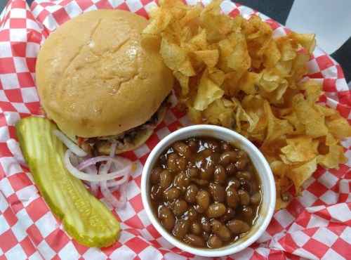 A hamburger on a bun with pickles and onions, served with a side of potato chips and baked beans in a red-checkered basket.