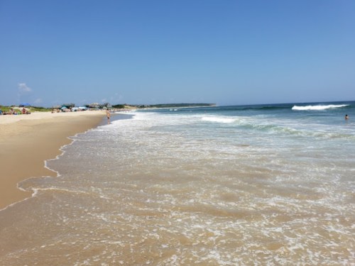 A sunny beach scene with gentle waves, sandy shore, and a few people enjoying the water and sun.