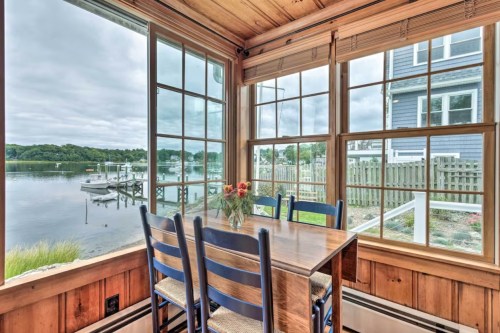 Cozy dining area with a wooden table and blue chairs, overlooking a serene waterfront view through large windows.