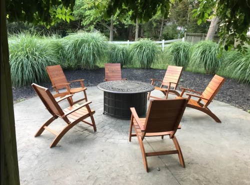 A cozy outdoor seating area with five wooden chairs arranged around a circular fire pit, surrounded by lush greenery.