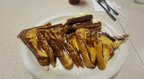 A plate of French toast drizzled with syrup, accompanied by several sausage links.