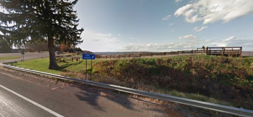Scenic overlook with benches and a sign, surrounded by grass and trees, under a clear blue sky.