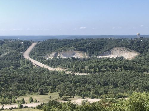 A panoramic view of a green landscape with a road, quarries, and distant water under a clear blue sky.
