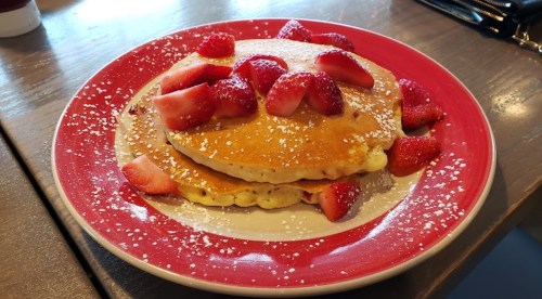 A stack of pancakes topped with fresh strawberries and powdered sugar, served on a red plate.