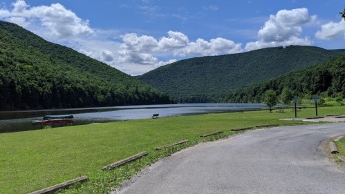 A serene lake surrounded by green mountains under a blue sky with fluffy clouds, featuring a grassy area and a path.