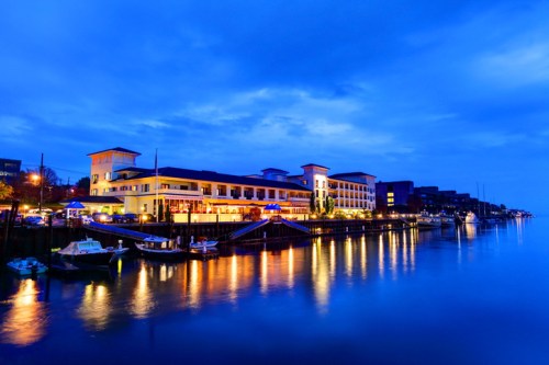 A serene waterfront scene at dusk, featuring illuminated buildings and boats reflecting on calm water.