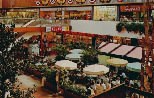 A bustling indoor mall with shops, dining areas, and people sitting under umbrellas in a vibrant atmosphere.