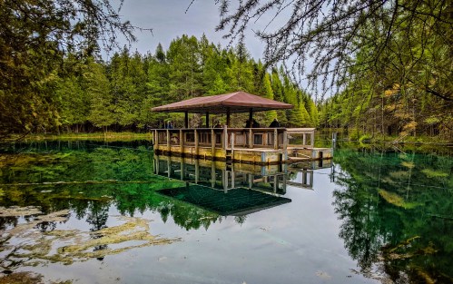 A wooden pavilion on stilts reflects in calm, clear water, surrounded by lush green trees.