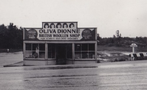 Black and white photo of Oliva Dionne British Woollen Shop, featuring a sign and a rainy road in front.
