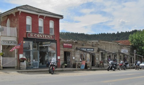Historic buildings line a street in a small town, featuring shops and parked motorcycles under a cloudy sky.