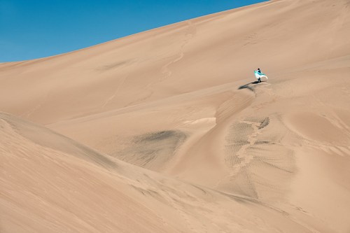 A person in a blue outfit stands on a vast, sandy dune under a clear blue sky.