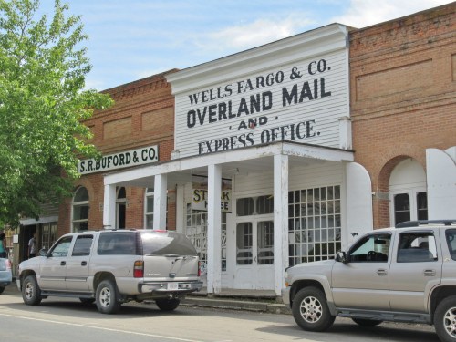 Historic building with "Wells Fargo & Co. Overland Mail and Express Office" sign, flanked by other shops and parked SUVs.