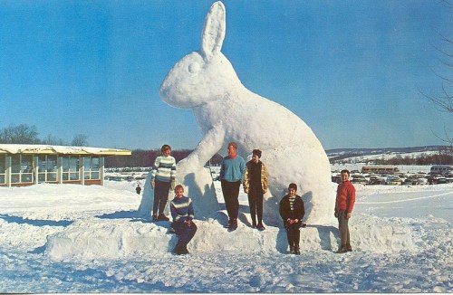 A group of people stands in front of a large snow rabbit sculpture in a snowy landscape.
