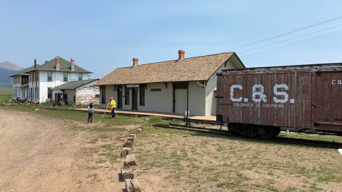 Historic train station with a wooden freight car nearby, surrounded by grassy land and a few people walking.