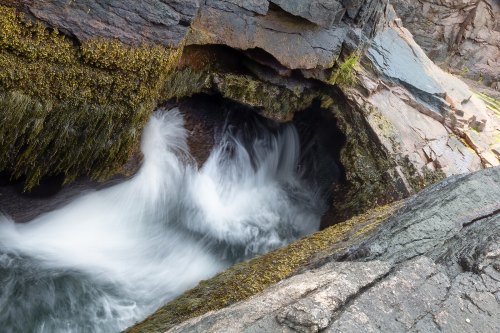 A rocky shoreline with flowing water creating a small whirlpool, surrounded by moss and textured stone.