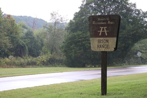 Sign for a bison range in a natural recreation area, surrounded by greenery and a road in the background.