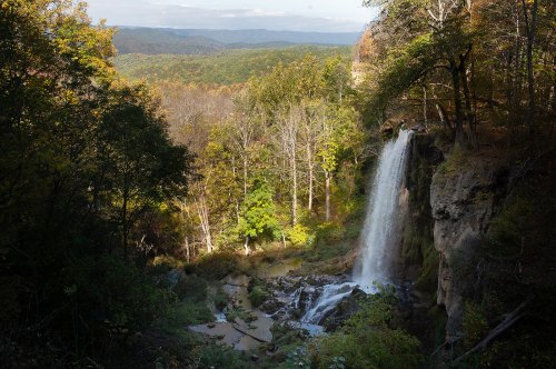 A scenic waterfall cascades down rocky cliffs, surrounded by lush green trees and rolling hills in the background.