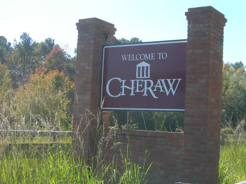 Welcome sign for Cheraw, framed by brick pillars and surrounded by tall grass and trees in the background.